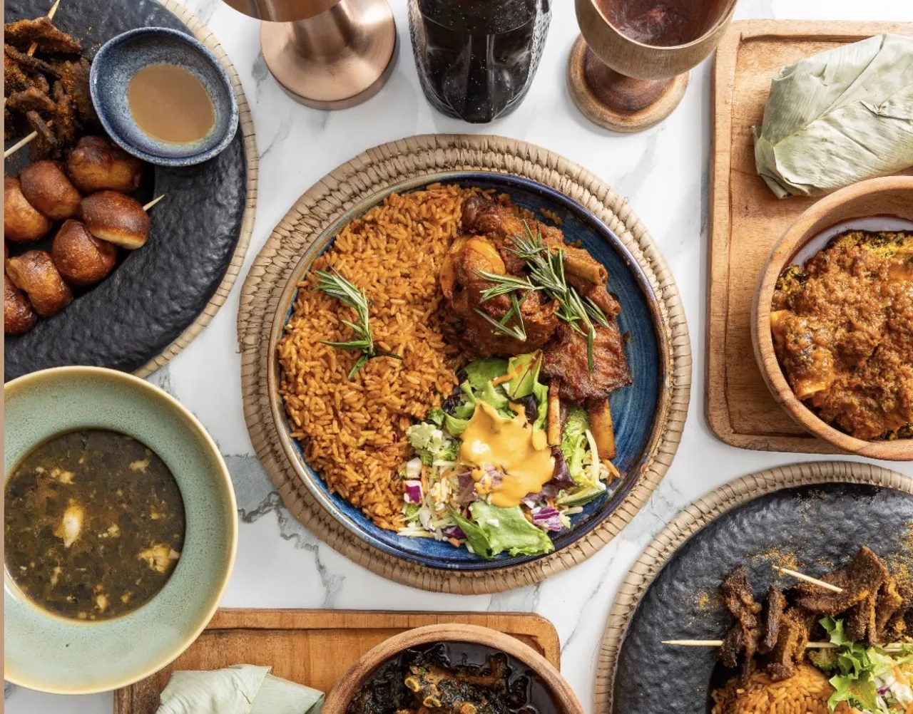An overhead spread of Ilé Yàn dishes including jollof rice, soups, suya and puff puff